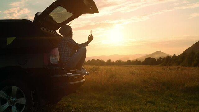 woman traveler holding smartphone taking photo landscape sitting car trunk looking on beautiful mountain landscape summer sky at sunset