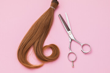 The cut off strand of female childrens hair of light brown color and scissors on a pink background, top view. Hairdressing services