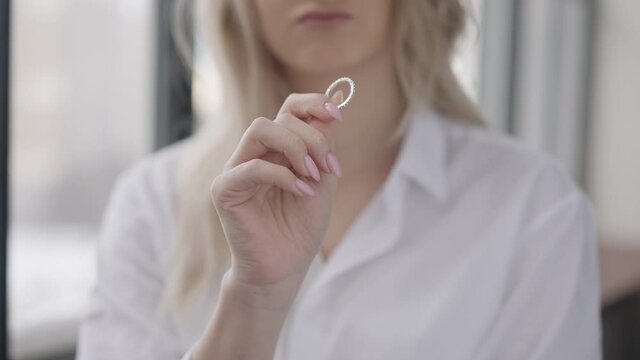 Close-up of ring in female Caucasian hand with blurred unrecognizable young woman sighing at background. Stressed cheated slim bride looking at jewelry thinking indoors. Heartbreak and divorce
