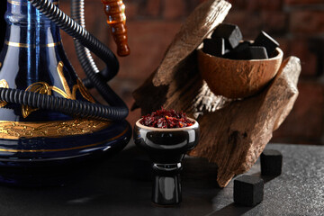 Obraz na płótnie Canvas Black glossy hookah bowl filled with apple shisha and prepared for smoking stands on a dark surface next to a beautiful hookah, charcoal and textured wood. Horizontal image.