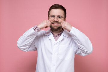 Funny doctor in a white coat makes the artificial smile on face with hands. Stress concept. Pink background.