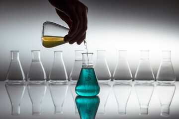 Hand pouring yellow in to blue liquid in scientific laboratory flask with glassware equipment on reflective surface.