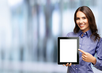 Happy smiling brunette businesswoman showing blank tablet pc monitor, with copy space area for some text, advertising or slogan, over blurred modern office interior background. Business woman indoors.