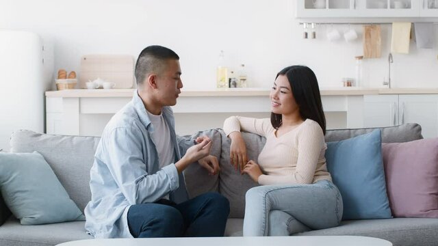 Cheerful Asian Couple Talking And Flirting Sitting On Couch Indoor