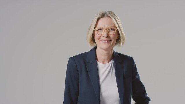 Studio portrait of smiling mature businesswoman wearing designer glasses against grey background in business suit looking at camera - shot in slow motion
