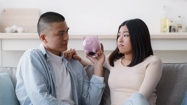 Unhappy Asian Couple Shaking Empty Piggybank Shrugging Shoulders At Home
