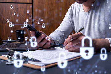 A businessman in casual wear signing the contract to prevent probability of risks in cyber security. Checking the phone. Padlock Hologram icons over the working desk.
