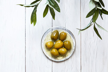 A bowl of pickled green olives. Minimal still life of an olive branch and a glass bowl of olives on...