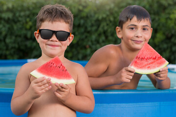 two cheerful boys by the blue pool are holding pieces of watermelon in their hands, summer