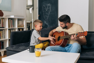 father do playing acoustic guitar with his son. they are sitting in living room