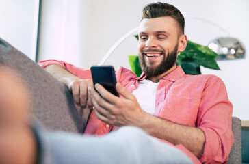 Young handsome happy bearded man in glasses is using smartphone for surfing in net, typing, and chatting, playing games, or working with some apps while relaxing on the couch at home