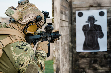 Soldier in the uniform (Cropat woodland pattern) taking aim at the target on the shooting range with his personal weapon. He is wearing ballistic helmet and plate carrier in coyote brown.