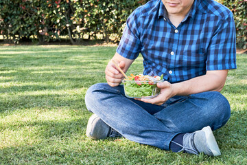 Young man sitting on the grass eating a fresh salad held in a glass bowl.