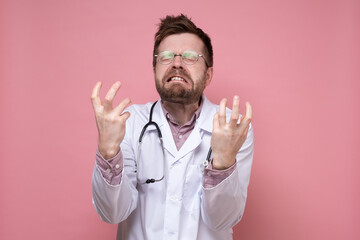 Doctor with a stethoscope around neck in stress, he bares teeth furiously and makes a gesture with hands. Pink background.