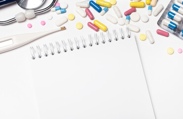 On a light background, multi-colored pills, a stethoscope, an electronic thermometer and a blank notebook with a place to insert text. Template. Medical concept.