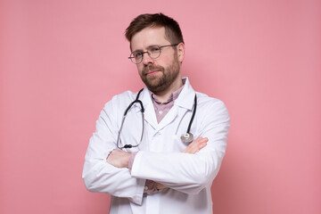 Calm, cute young doctor with a stethoscope around neck looks confidently into the camera with crossed arms. Pink background.