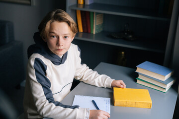 High-angle view of clever pupil boy sitting at desk with paper workbook and preparation to doing homework in children room, looking at camera. Child schoolboy studying at home near window.