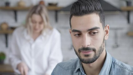 Headshot portrait of young handsome bored man listening to blurred angry woman arguing at background. Close-up of Middle Eastern husband listening Caucasian wife shouting and gesturing