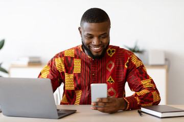 Positive Black Man In Traditional African Costume Messaging On Smartphone At Workplace