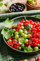 Green gooseberries and red currants with leaves in a green ceramic plate.