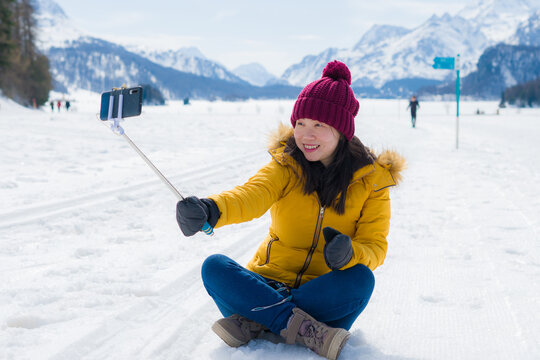Winter holidays selfie in the snow - young happy and cheerful Asian Korean woman on taking self portrait with mobile phone in cold snowy mountain at Swiss Alps