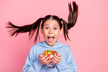 Photo portrait of shocked schoolgirl keeping glass with sweet candies staring with flying hair isolated on pastel pink color background