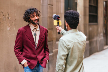 young caucasian trendy man standing in the street doing a live event. He is recorded by a latin...