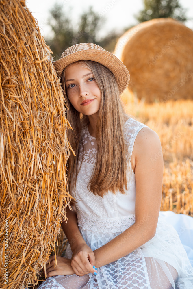 Canvas Prints Beautiful young girl in a white dress and a straw hat sitting near the bale of straw. Outdoor portrait of a positive female