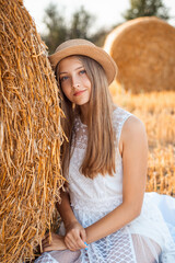 Beautiful young girl in a white dress and a straw hat sitting near the bale of straw. Outdoor portrait of a positive female