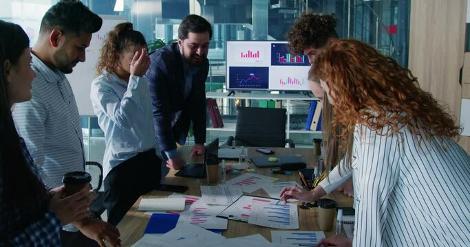 Diverse coworkers having corporate meeting, standing around conference table and analyzing financial papers. Business students engaged in teamwork with male mentor.