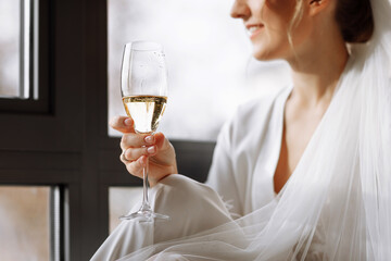 Bride s morning. Bride drinking champagne in the peignoir. young woman is sitting on a large window in a hotel room in bathroom. Beautiful girl in white wedding robe. wedding day