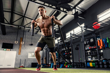 Fototapeta na wymiar A muscular man without a shirt pulls a rope in a modern gym with equipment. A strong sportsman full of energy prepares to exercise in functional training. Getting ready for the fitness challenge