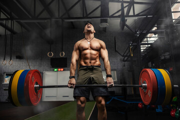 Fototapeta na wymiar Weightlifter in the gym, a moment before a strong movement. Strong muscular man holds a heavy barbell in his hands and performs a dead lift in a gym with a dark atmosphere. Screaming during exercises
