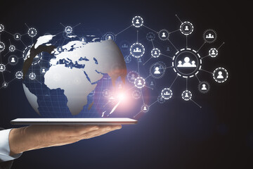 Fototapeta premium Hand with a tablet holding the globe icon with muptiple small application icons near it on a dark background. Worldwide web and social communication concept