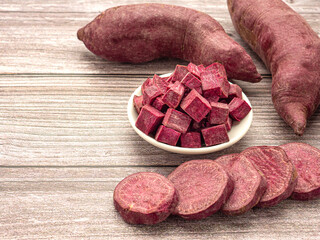 A pile of purple yams whole, sliced square in a bowl and sliced circle placed on a wooden table with copy space for text. Purple sweet potatoes. Top view. Vegan fruit and healthy fruits concept