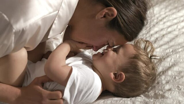 Authentic close up shot of young happy mother is tickling and playing with her toddler baby boy in a nursery woke up in a morning. Concept of love, parenthood, childhood, life, maternity, motherhood.