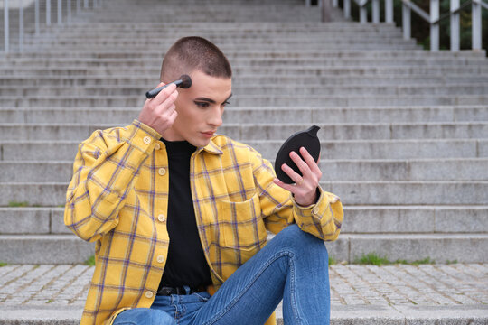 Young non binary gender person applying make up with a brush in front of a mirror. Androgynous lifestyle.