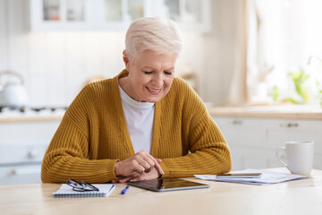 Positive grey-haired senior woman using digital tablet in kitchen
