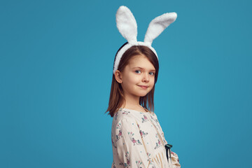 Pretty little girl in white shirt and bunny ears posing adorably on blue background smiling at...