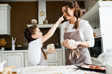 A sweet daughter touches her finger in flour to the nose of her mother who sculpts cookies from the dough