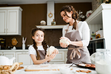 Mom in an apron teaches her little daughter how to sculpt from raw dough, shaping baking blanks with her hands