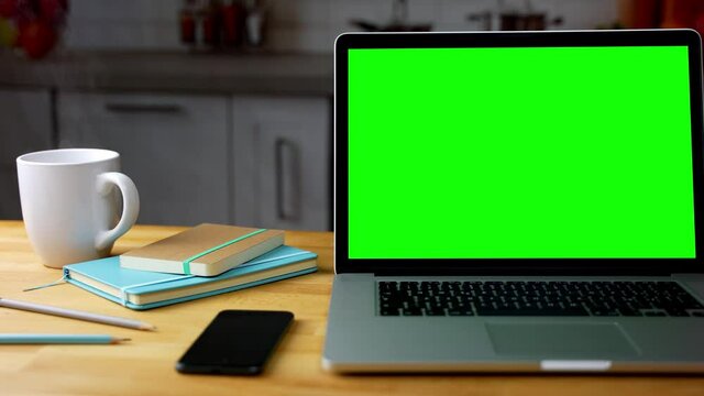 Laptop in green screen at home on a wooden table, with a mobile phone, pencils a cup of coffee and blue notepads, notebooks. Distance learning and working.