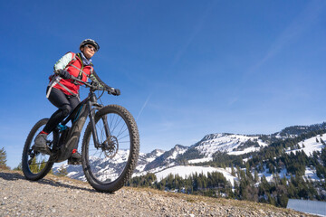 Fototapeta na wymiar senior woman mountainbiking below the Nagelfluh mountain chain with Hochgrat summit on a e-mountainbike in early spring, in the Allgaeu Area near Steibis and Obers, a part of the bavarian alps,Germany