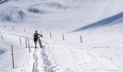 Fototapeta na wymiar senior woman mountainbiking and pushing her e-mountainbike through a snow spot in early spring, in the Allgaeu Area, a part of the bavarian alps,Germany