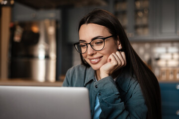 White smiling woman in eyeglasses working with laptop at home kitchen