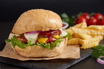 Homemade hamburger or burger with fresh vegetables and cheese lettuce and mayonnaise served, French fries on pieces of brown paper on black stone table . concept of fast food and junk food