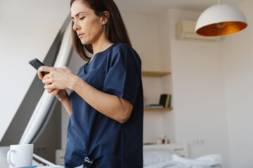 Puzzled woman doctor using mobile phone while standing at home
