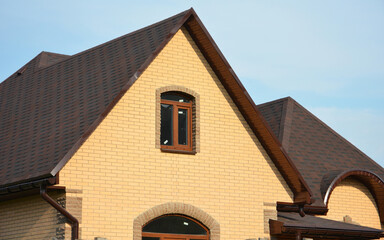 A brick house construction with a brown asphalt shingled roof, attic window, roof gutters and soffit. A complex roof with asphalt roof shingles.