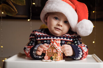 Little baby boy with wearing Santa hat playing with toy gingerbread house sitting in baby high chair at home. Funny baby, Christmas celebration concept.