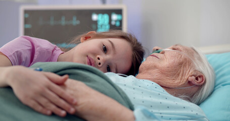 Close up of sad kid hugging sleeping sick grandmother lying together in hospital bed
